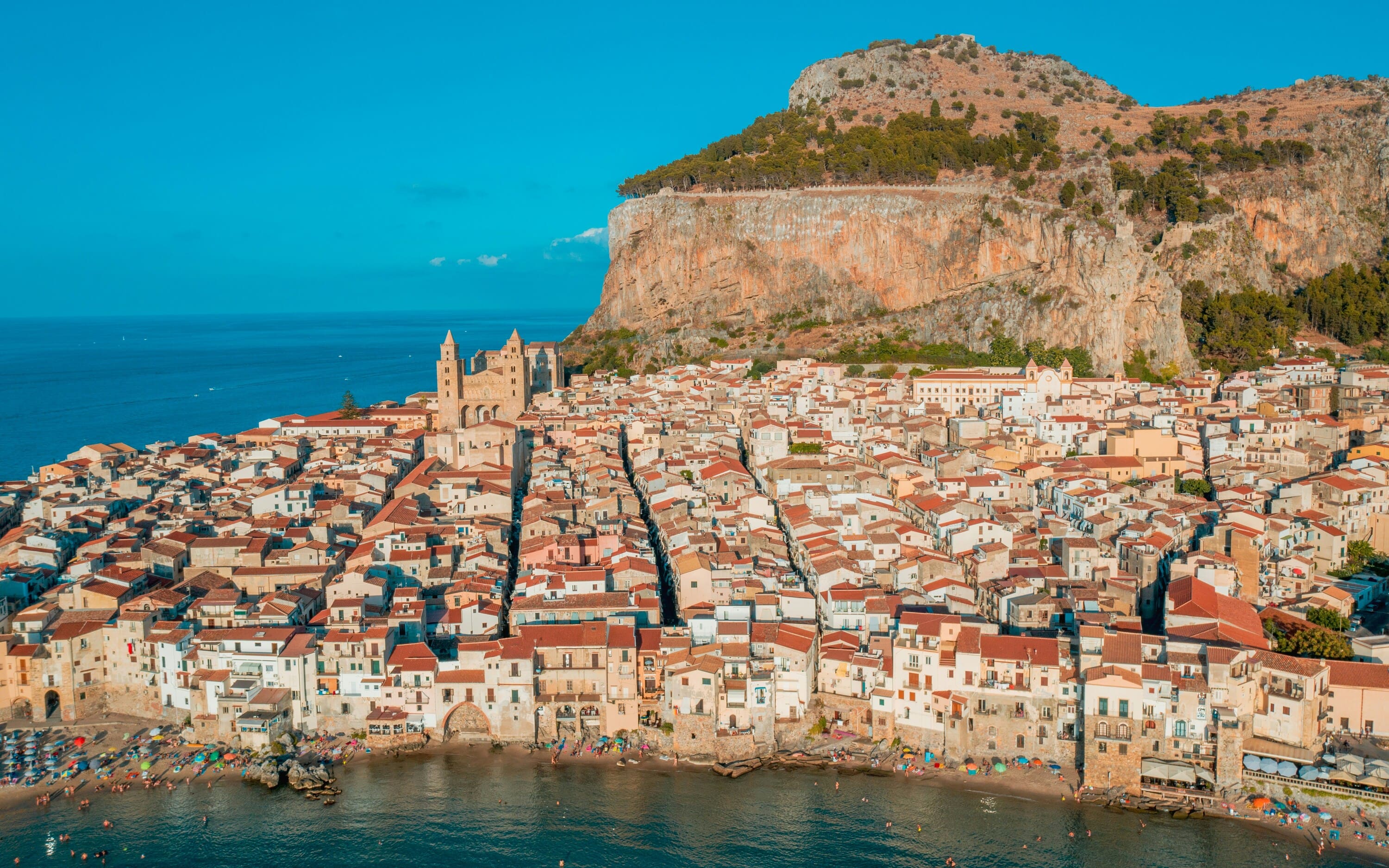 Aerial view of a coastal hillside town with dense white and beige buildings, terracotta roofs, a church tower, and a large rocky cliff rising behind it under a clear blue sky.