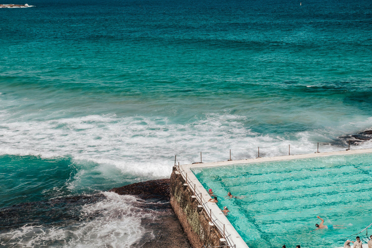A high-angle view of a coastal ocean pool next to the turquoise sea, with waves crashing against the shore and swimmers visible in the pool.