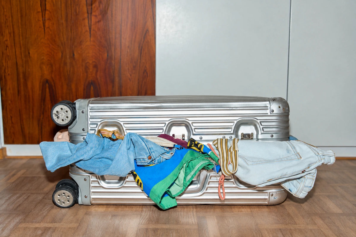 Silver hard-shell suitcase overflowing with colorful clothes, denim jeans, and shirts on a wooden floor.